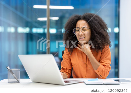 An office worker holds her throat in discomfort while sitting at a desk with a laptop. She appears to be dealing with a health issue during work hours, looking visibly uncomfortable. An office worker holds her throat in discomfort while sitting at a desk with a laptop. She appears to be dealing with a health issue during work hours, looking visibly uncomfortable. 120992845