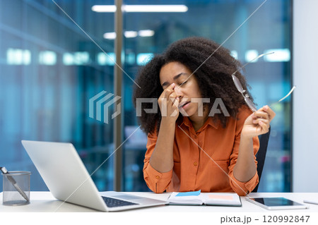 Young woman businesswoman appears stressed and tired at her desk in a modern office setting. She holds her glasses while using a laptop, symbolizing fatigue and work pressure. Young woman businesswoman appears stressed and tired at her desk in a modern office setting. She holds her glasses while using a laptop, symbolizing fatigue and work pressure. 120992847