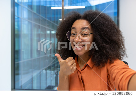 Young woman businesswoman in an orange shirt smiling and giving a thumbs up. She is in a bright, modern office space, conveying positivity and confidence. 120992866