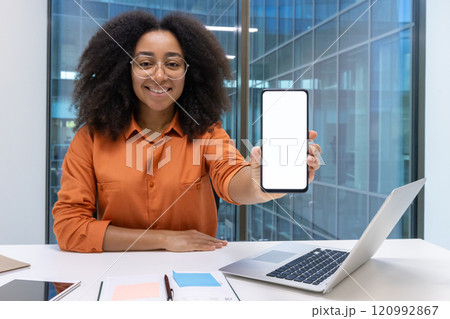A cheerful woman in an office setting shows a smartphone with a blank white screen. The modern work environment includes a laptop, notebook, and window view. 120992867