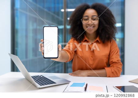 Young woman businesswoman displaying a smartphone at her desk in a modern office environment. She is wearing glasses and smiling, creating a professional yet approachable atmosphere. 120992868