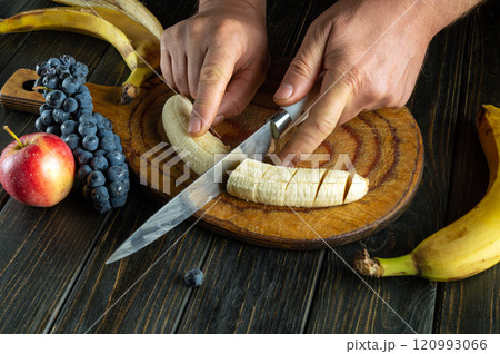 Hands skillfully slice ripe bananas on a rustic wooden board, surrounded by fresh blueberries, black grapes, and a crisp red apple, creating a vibrant and inviting fruit platter Hands skillfully slice ripe bananas on a rustic wooden board, surrounded by fresh blueberries, black grapes, and a crisp red apple, creating a vibrant and inviting fruit platter 120993066