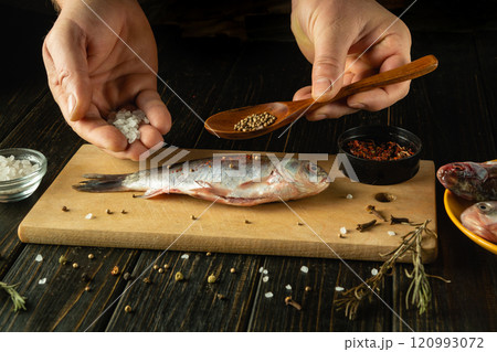 Hands carefully season a fresh fish on a wooden board using salt, herbs, and spices, creating an aromatic preparation for a delicious meal. The kitchen exudes warmth and creativity. 120993072