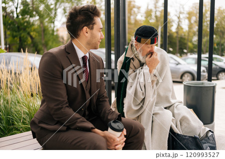 Compassionate entrepreneur with coffee engaging in conversation with homeless senior citizen. Kind businessman in suit listening with empathy to homeless elderly man talking sitting on urban bench. 120993527
