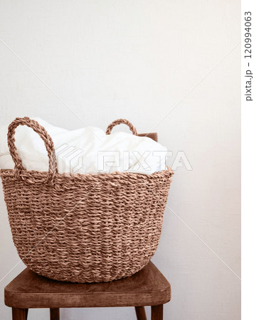 A natural woven basket with white linens inside, placed on a rustic wooden chair against a neutral white wall. A natural woven basket with white linens inside, placed on a rustic wooden chair against a neutral white wall. 120994063