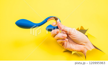Female hand holding blue vibrator sticking out of yellow paper background. Copy space. 120995963