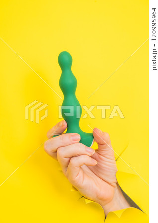 Female hand with green anal plug sticking out of yellow paper background. 120995964