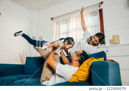 Family fun takes flight in the living room with an airplane game. The child held high on the father's shoulders and supported by the mother enjoys a cheerful carefree time. 121000070