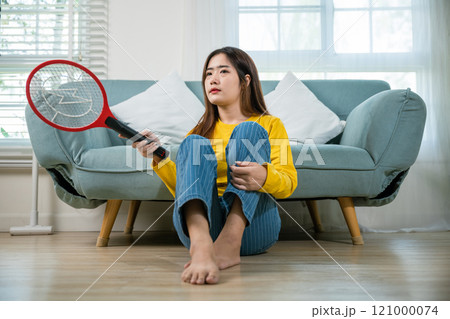 Asian female sitting floor using mosquito device swatter or electric net racket, Young woman killing mosquitoes hand holding fly swatter like weapon in living room at home, technology insect killer 121000074