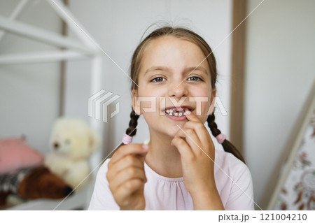 Little Caucasian girl smiling and holding first lost baby tooth in cozy home environment showcasing childhood milestone. 121004210