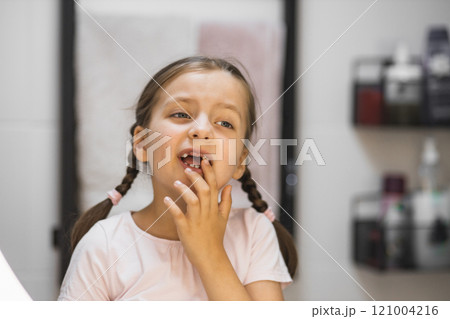 Caucasian girl with braided hair examines her loose tooth and missing teeth in bathroom mirror. Young child with tooth gap experiences curiosity and wonder during dental milestone. 121004216