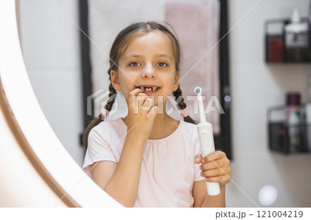 Little Caucasian girl with missing baby teeth using electric toothbrush and smiling in bathroom mirror, showing confidence and dental care. 121004219