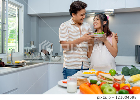 Asian man stand beside pregnant woman and hold the dish of food and look to each other during woman prepare to cook in the kitchen of their house. 121004698