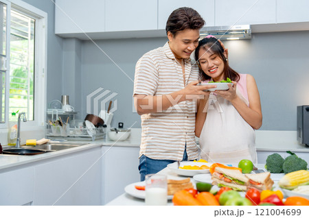 Asian man stand beside pregnant woman and hold the dish of food during woman prepare to cook in the kitchen of their house. 121004699