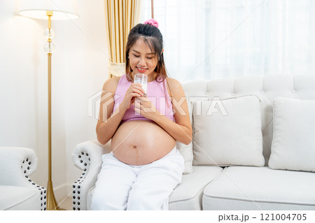 Asian pregnant woman sit on sofa and hold a glass of milk and touch her belly also look at the glass with happiness. Asian pregnant woman sit on sofa and hold a glass of milk and touch her belly also look at the glass with happiness. 121004705