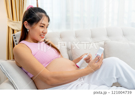 Side view of Asian pregnant woman lie on sofa and use tools for listen sound in her belly and look happy at home. Side view of Asian pregnant woman lie on sofa and use tools for listen sound in her belly and look happy at home. 121004739