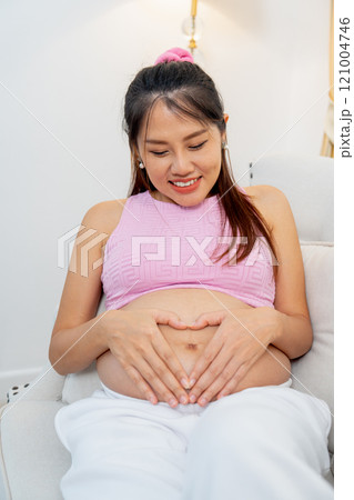 Vertical image of Asian pregnant woman sit on sofa and put hands on her belly with heart shape and smiling. Vertical image of Asian pregnant woman sit on sofa and put hands on her belly with heart shape and smiling. 121004746