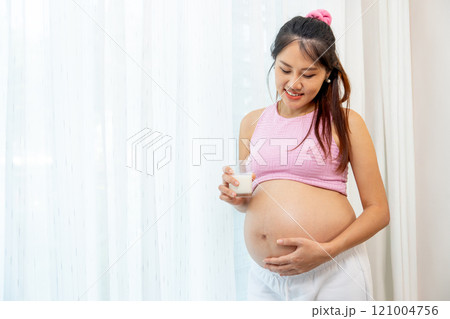 Asian pregnant woman stand with hold a glass of milk and stand in front of white curtain also look at her belly with happiness smile. Asian pregnant woman stand with hold a glass of milk and stand in front of white curtain also look at her belly with happiness smile. 121004756