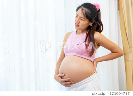 Asian pregnant woman stand in front of white curtain and action like she feel backpain that she touch her back and belly. 121004757