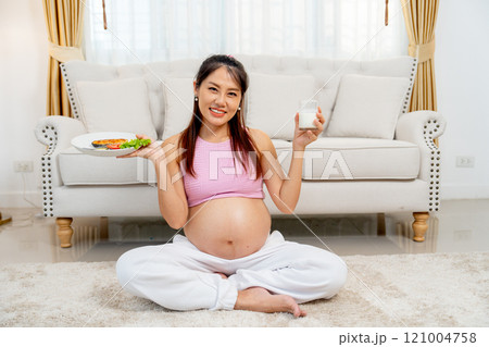 Asian pregnant mother sit on the floor of living room with holding disk of food and milk and look at camera with smiling. 121004758