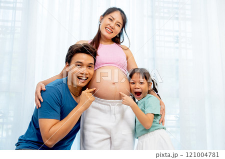 Asian father and little daughter hug and point to belly of pregnant mother and express happiness with smiling in front of white curtain. 121004781