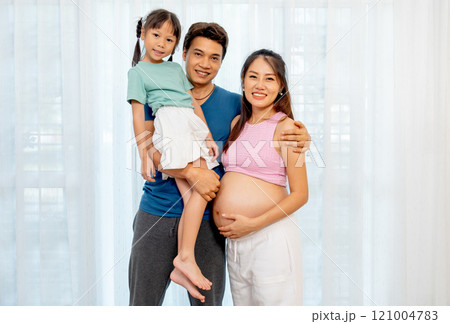 Asian father carry little girl and stand beside pregnant mother in front of white curtain with smiling and look at camera. Asian father carry little girl and stand beside pregnant mother in front of white curtain with smiling and look at camera. 121004783