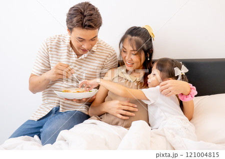 Asian father hold dish of food and little touch food to express hungry and stay beside pregnant mother on bed in concept of happiness family in their house. 121004815