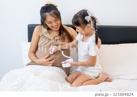 High view of little Asian girl enjoy to play with sound check tool on belly of pregnant mother and stay on bed of their house to express love and care in family. 121004824