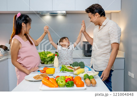 Little Asian girl touch hands together with father and pregnant mother and stay in kitchen to prepare for cooking with happiness. Little Asian girl touch hands together with father and pregnant mother and stay in kitchen to prepare for cooking with happiness. 121004848