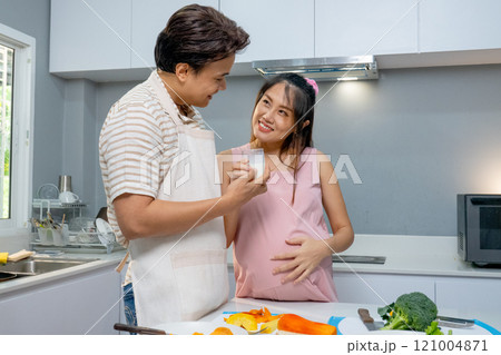 Asian man hold a glass of milk give to pregnant woman and look each other with love and care stay in the kitchen of their house. 121004871