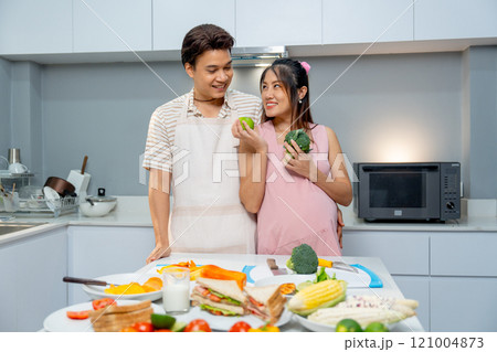 Asian man stand beside pregnant woman who hold apple and vegetable and express love and care stay in the kitchen of their house. Asian man stand beside pregnant woman who hold apple and vegetable and express love and care stay in the kitchen of their house. 121004873