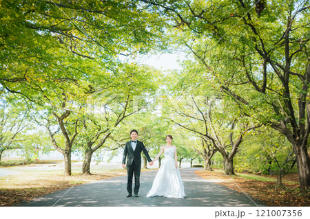 公園の並木道を手を繋いで歩くドレス姿のカップル（ウェディング・ウェディングフォト・新郎新婦） 121007356