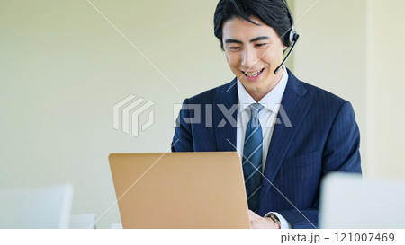 ノートパソコンを見ながらヘッドセットで通話する男性ビジネスパーソン ノートパソコンを見ながらヘッドセットで通話する男性ビジネスパーソン 121007469