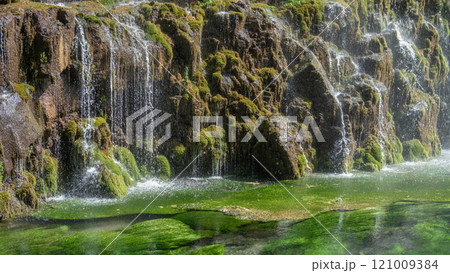 A serene waterfall cascades over moss-covered rocks into a clear, green mountain river. 121009384