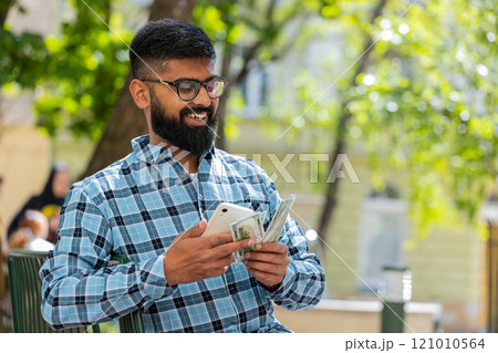 Happy Indian man counting money dollar cash, use smartphone calculator app in urban city street Happy Indian man counting money dollar cash, use smartphone calculator app in urban city street 121010564
