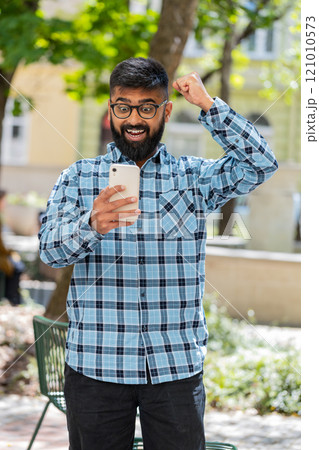 Indian man use mobile smartphone celebrating win good message news outdoors on urban city street Indian man use mobile smartphone celebrating win good message news outdoors on urban city street 121010573