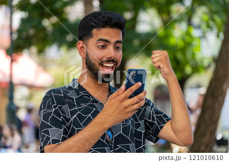 Excited happy joyful winner Indian man using smartphone celebrating win good message news outdoors 121010610