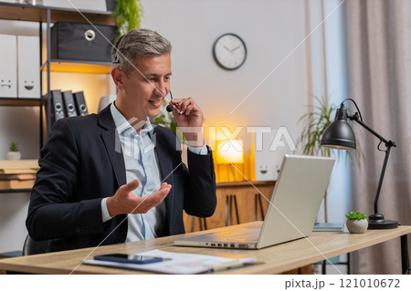 Confident young businessman in headset talking on video call using laptop computer at office desk 121010672