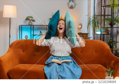 Irritated young woman reading book cover ears with pillows annoyed by loud noise sitting on sofa 121010684