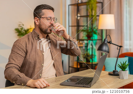 Focused Caucasian businessman sitting at desk drinking water while working on laptop in home office 121010756