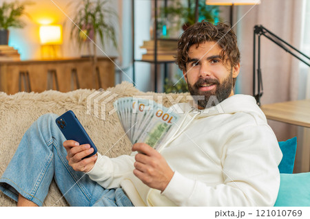 Successful rich young Indian man holding dollar bill and smartphone sitting on sofa couch at home 121010769