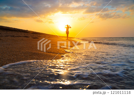Slender Caucasian young woman in a hat walks along the seashore or ocean at sunset. Low angle water close wide angle 121011118