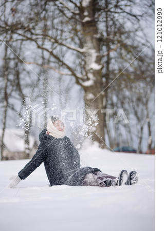 Young woman enjoying wintertime, sitting in snow and throwing it in the air in a park Young woman enjoying wintertime, sitting in snow and throwing it in the air in a park 121012990