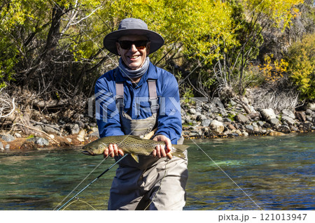 Portrait of smiling fly-fisherman holding native brown trout from Traful River before releasing it unharmed 121013947