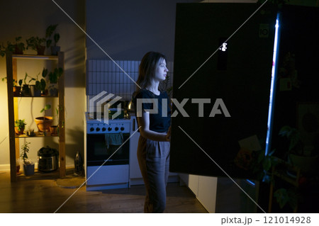 Woman standing in front of open refrigerator at night Woman standing in front of open refrigerator at night 121014928