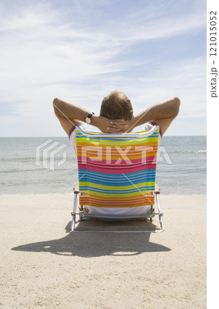 Rear view of man relaxing on deck chair on sandy beach 121015052