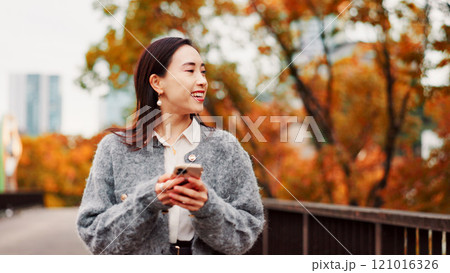 Thinking, phone and happy woman at park for travel, text or message on app in Japan. Smile, mobile and Asian person outdoor for dream, planning vacation and post on social media profile on internet 121016326