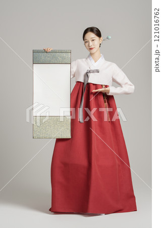 A woman in her 20s wearing a hanbok, wearing a colorful hairpin, and posing while holding a hanging scroll. A woman in her 20s wearing a hanbok, wearing a colorful hairpin, and posing while holding a hanging scroll. 121016762