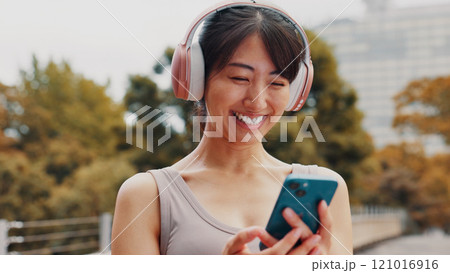 Smile, woman and headphones with phone in city of listening to music, fitness podcast and social media. Athlete, mobile app and streaming radio for exercise routine, training website and Japan bridge 121016916