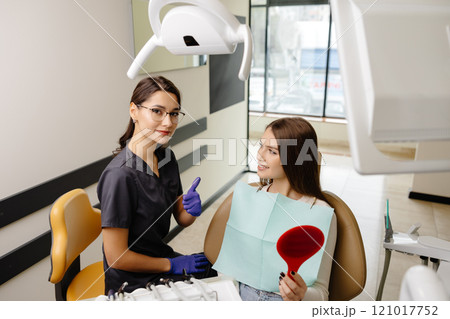 Woman is happy with results after oral hygiene and teeth whitening at dentist office. Patient is smiling and looking at the mirror Woman is happy with results after oral hygiene and teeth whitening at dentist office. Patient is smiling and looking at the mirror 121017752
